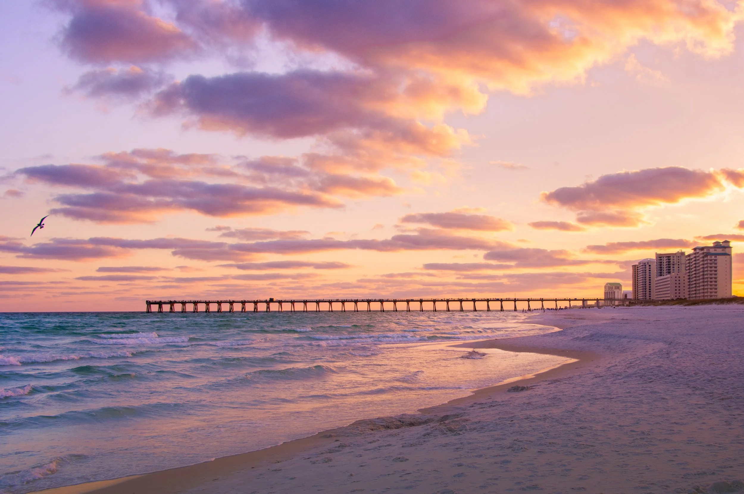 Sunset over a beach with pink and purple clouds, a pier extending into the ocean, and high-rise buildings in the distance.