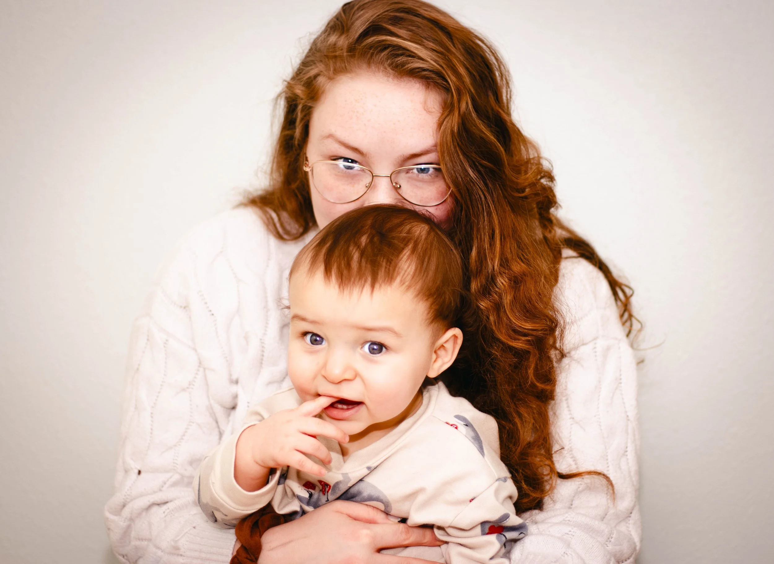 A woman with red hair and glasses holding a young child with red hair against a plain background.