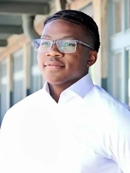 Young man wearing glasses and a white shirt, standing outdoors near a building with large windows.