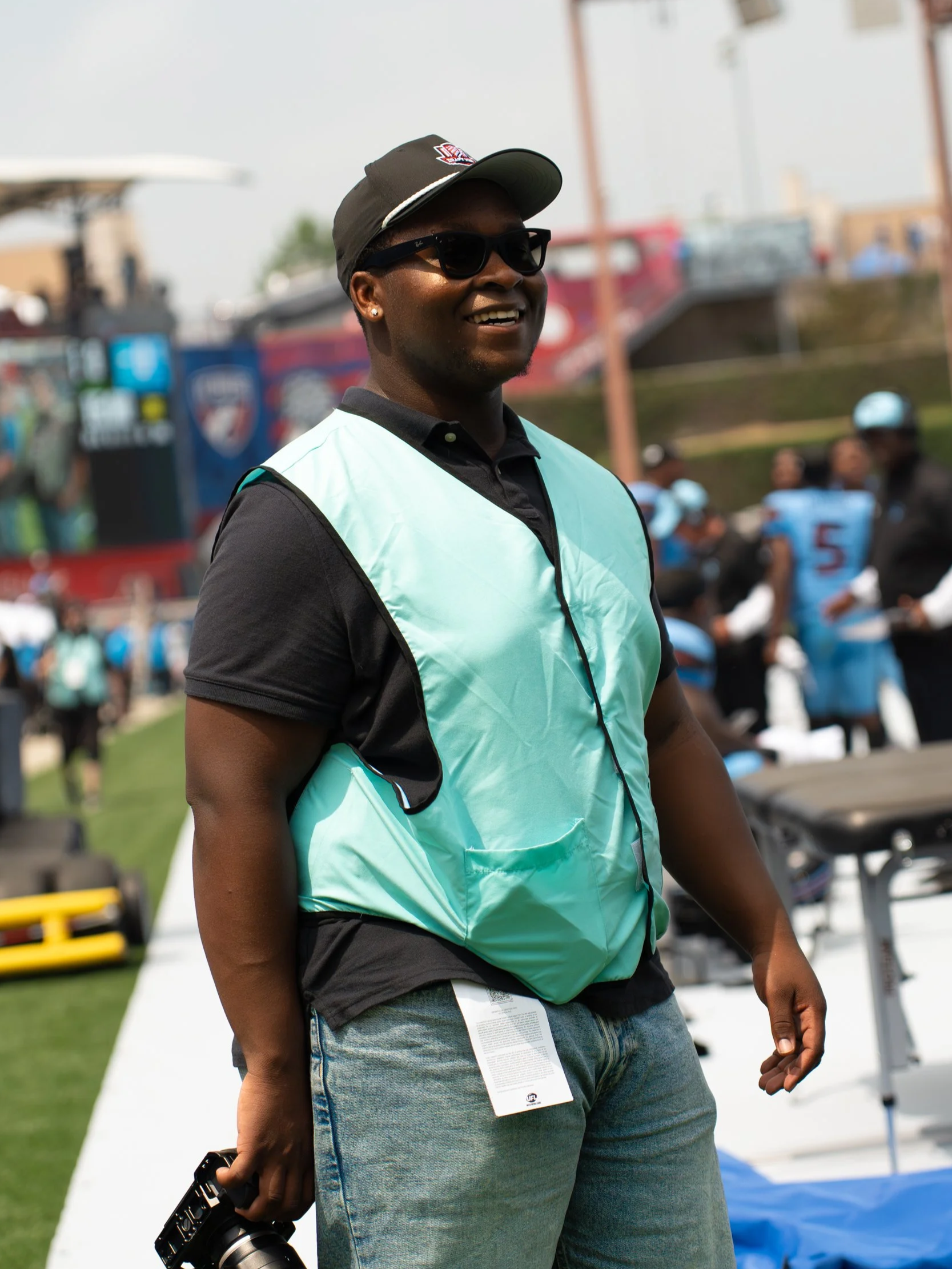 A smiling man with dark sunglasses and a black cap, wearing a black shirt and a teal vest, at an outdoor event with people and equipment in the background.