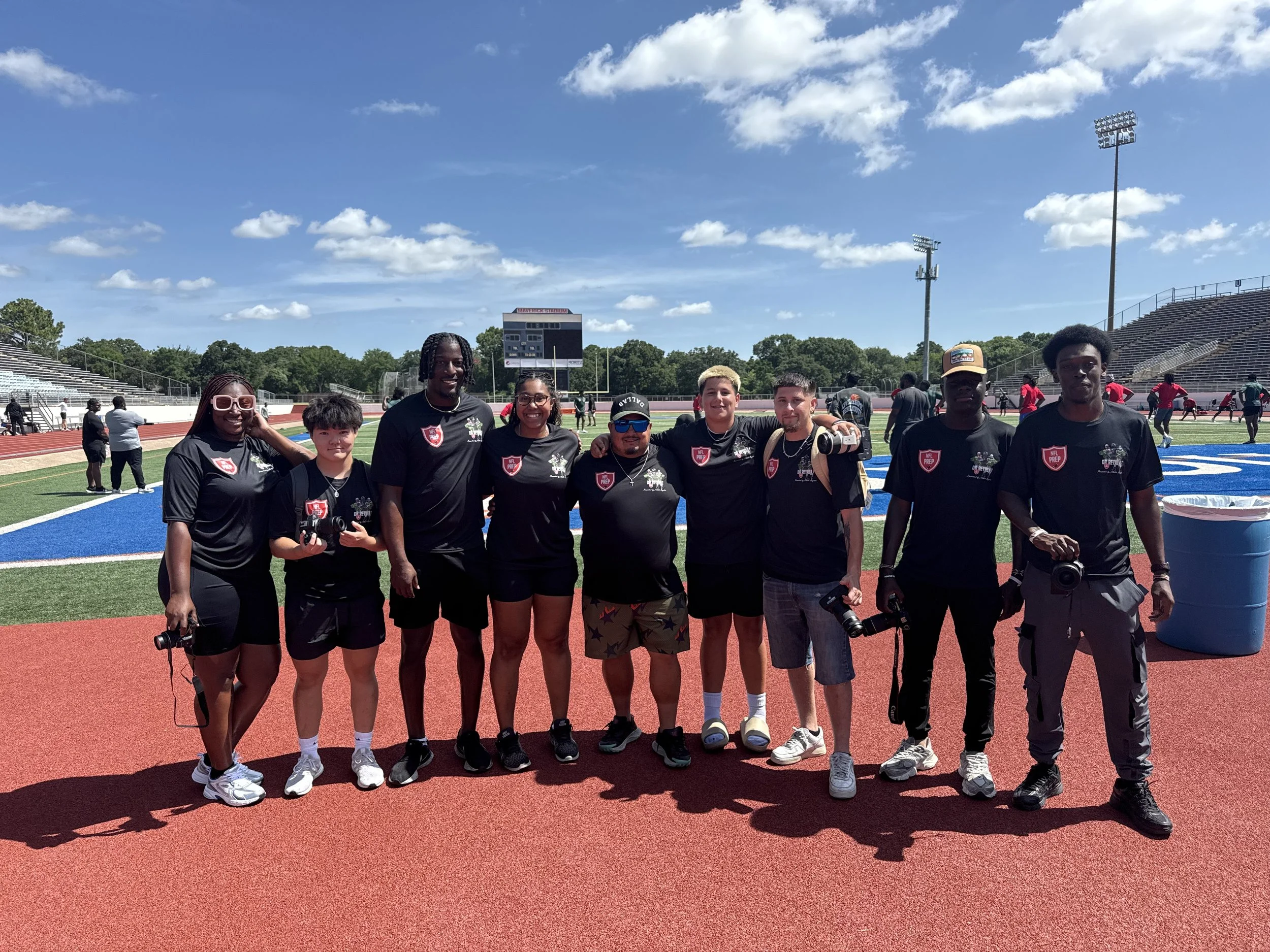 A group of nine young people standing on a track field at a stadium, smiling at the camera, with a digital scoreboard and stadium seating in the background.
