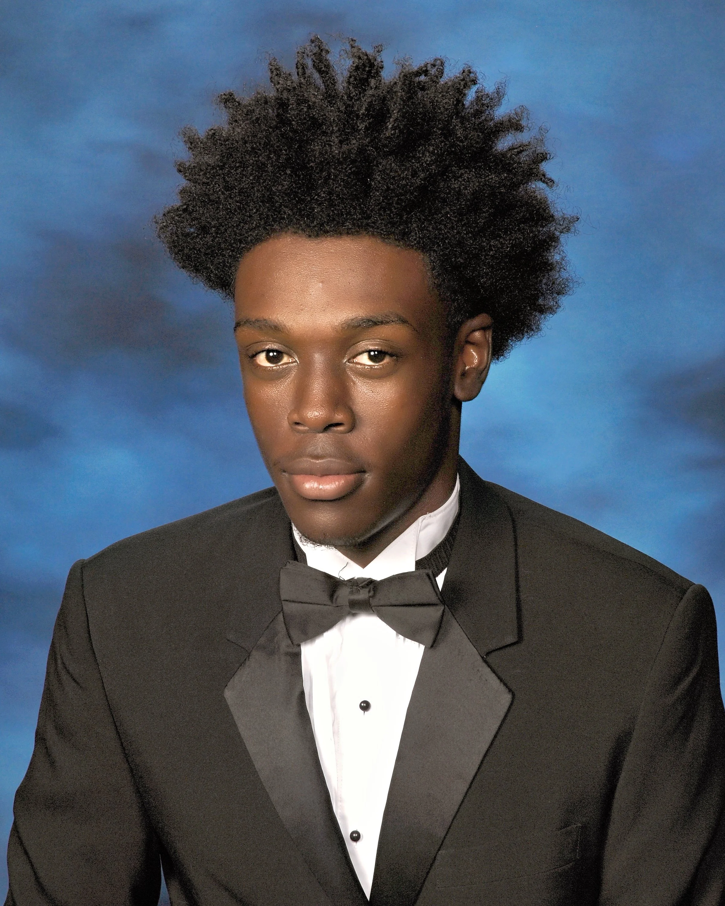 Close-up of a person with dark skin and natural curly hair wearing a black tuxedo with a white shirt and black bow tie, against a blue background.