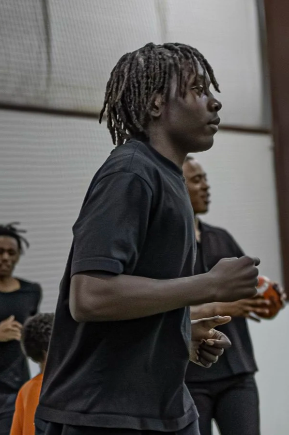 Young man with dreadlocks in a black shirt standing indoors, with other people in the background.
