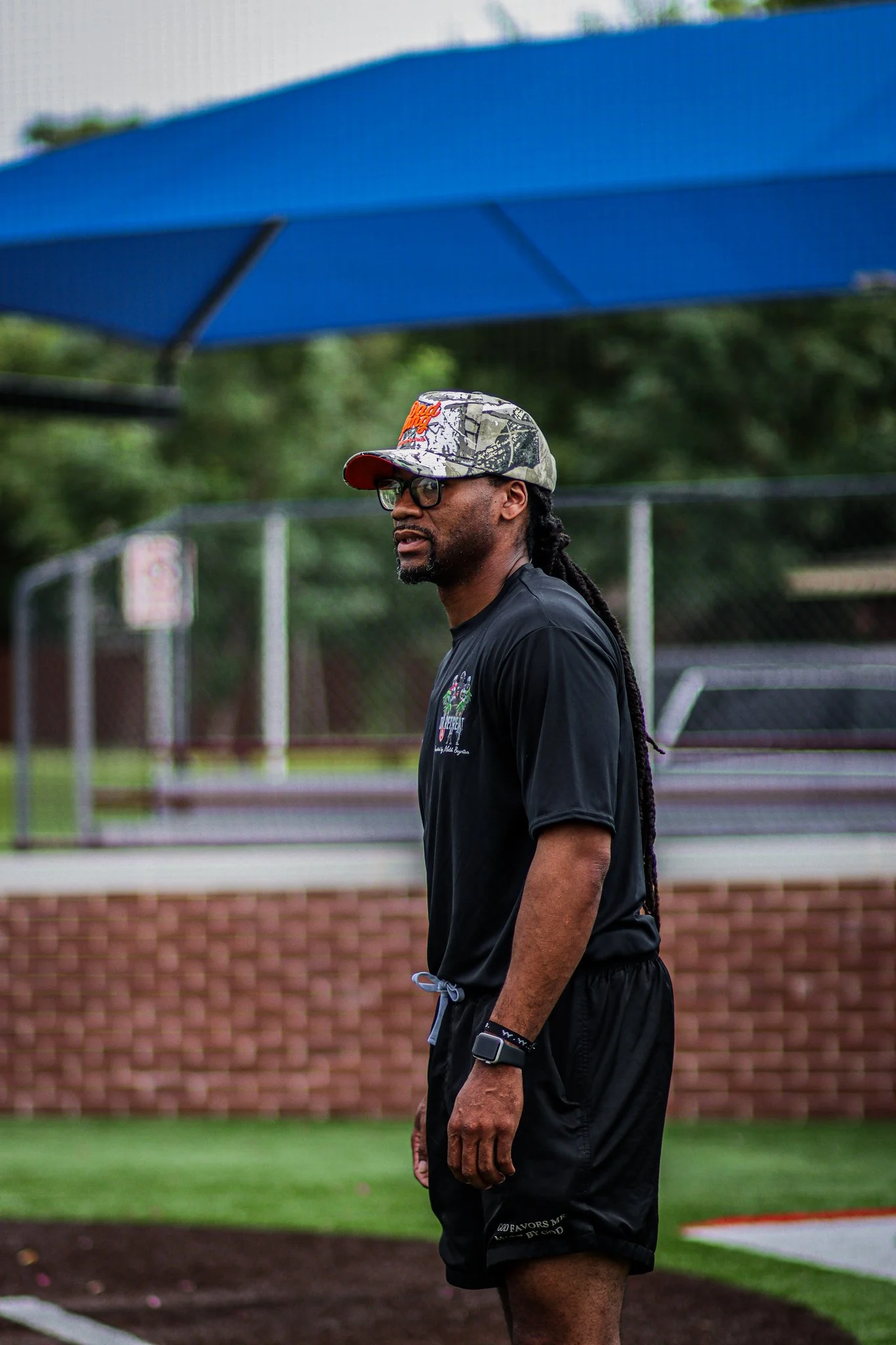 A man with dreadlocks, glasses, and tattoos on his arm standing on a sports field. He is wearing a camouflage cap and black athletic clothing, and is looking to the side with a focused expression.