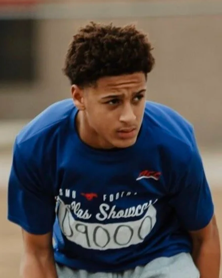 Young male athlete with curly hair wearing a blue shirt with a race bib, at a track and field event.