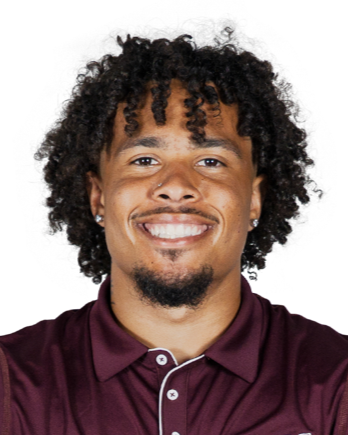 A young man with curly hair and a goatee, smiling, wearing a maroon polo shirt.