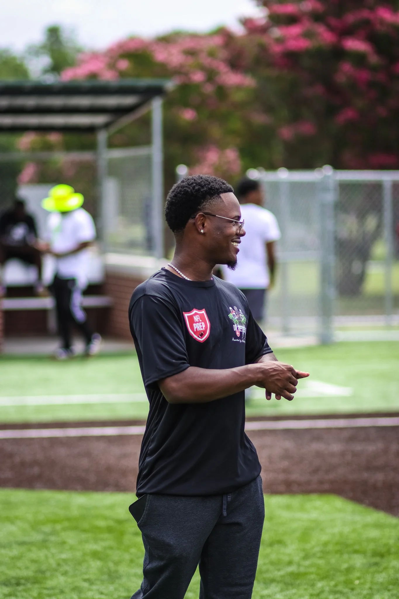 A smiling young man with glasses, wearing a black NFL Prep shirt and dark pants, standing outdoors on a sports field with a chain-link fence and pink trees in the background.