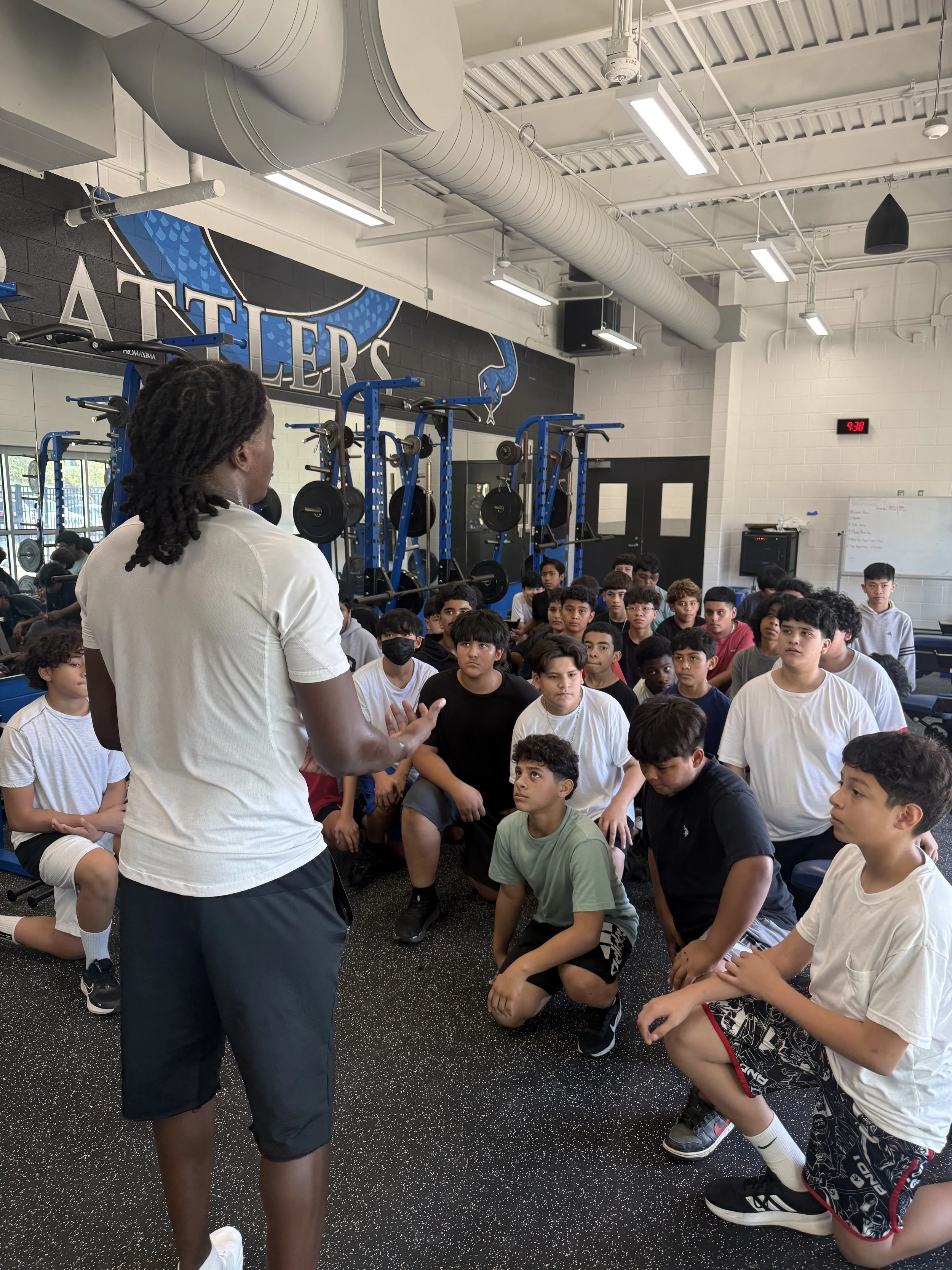 A coach or trainer standing in front of a group of young male athletes who are kneeling and sitting on black rubber gym flooring in a weight room with blue and black equipment. The room has high ceilings with exposed ductwork and a large sign that says 'ATHLETES' on the wall behind the group.