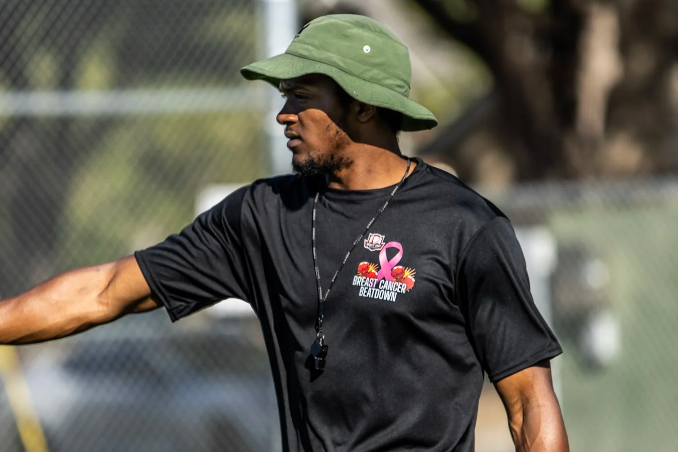A man wearing a green bucket hat and a black T-shirt with a breast cancer awareness ribbon and logo, standing outdoors near a chain-link fence.