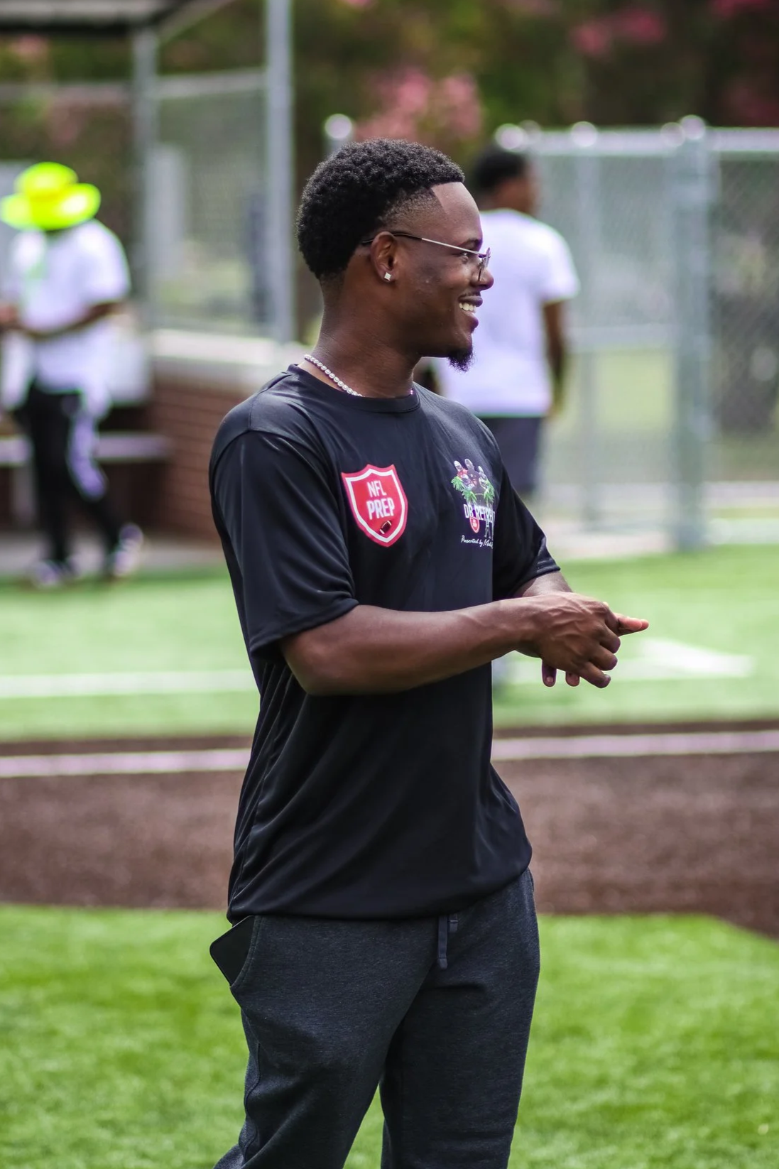 A young man wearing glasses, a black t-shirt with NFL PREP logo, and a necklace, standing on a sports field, smiling and looking to the side.