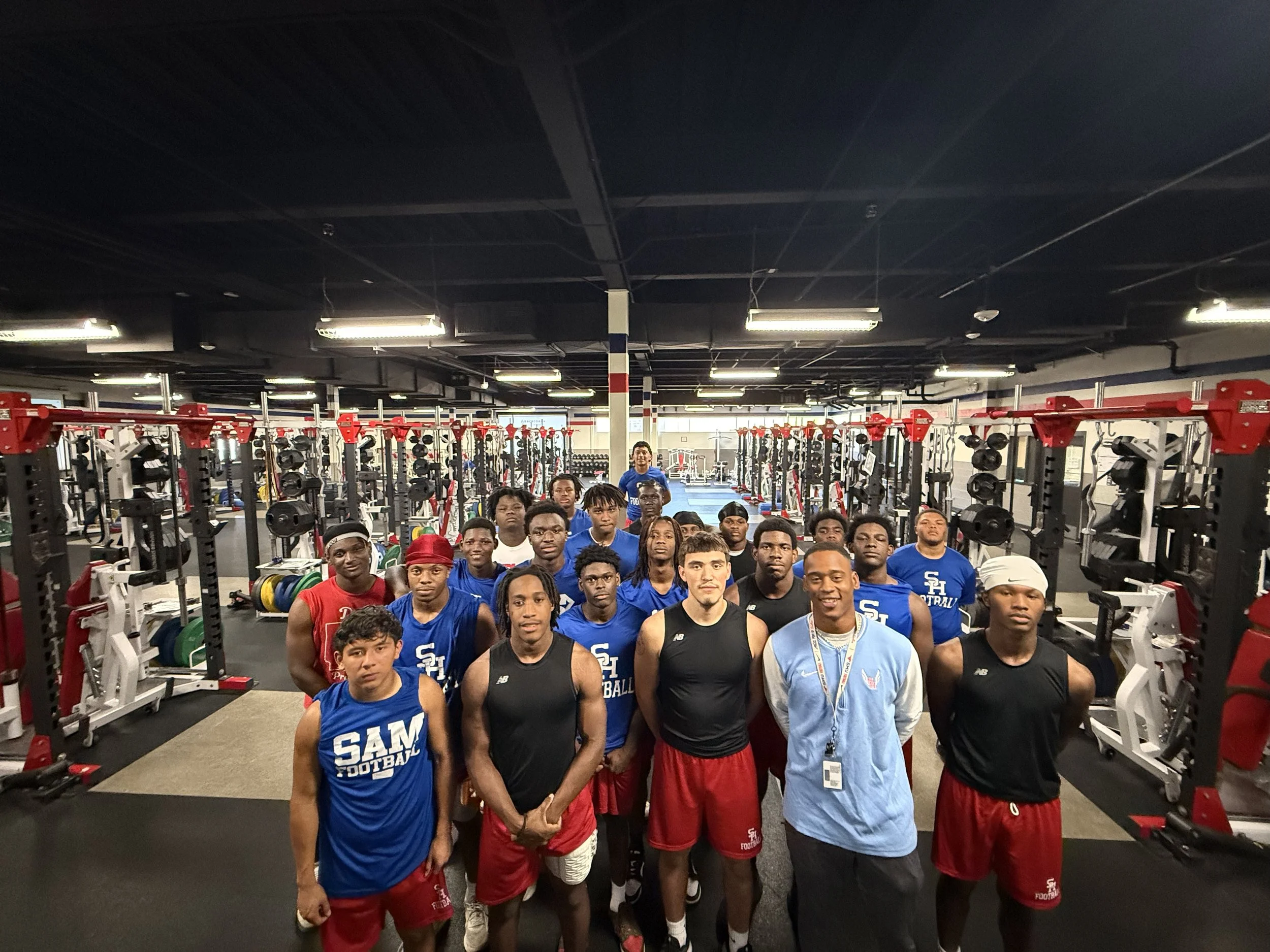 Group of young men in athletic clothing standing together inside a gym, posing for a photo, with gym equipment in the background.