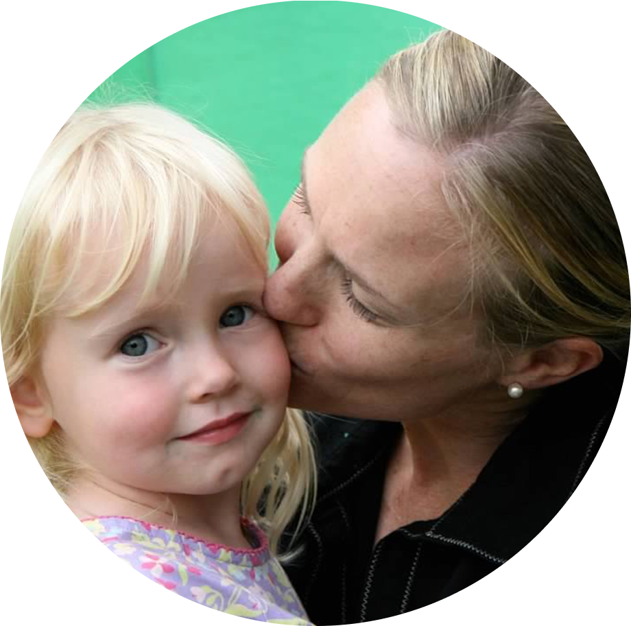 A woman with blonde hair sitting on a chair, smiling, wearing a sleeveless black top and dark jeans, against a plain white background.