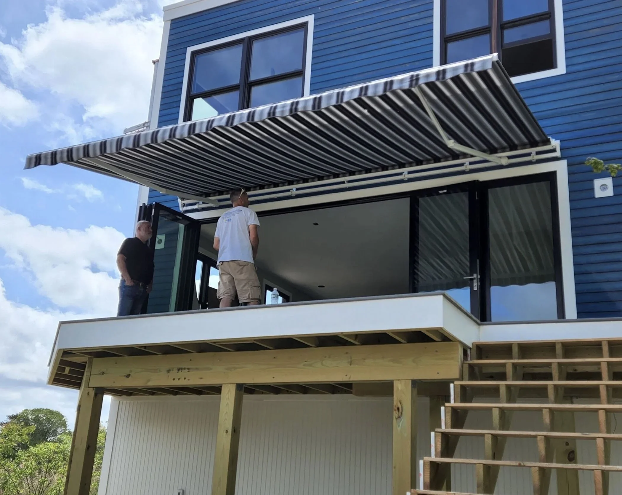 Construction workers installing a retractable awning on a modern blue house with an upstairs balcony. The house has large windows and a sliding glass door leading to the balcony. The balcony is elevated with wooden supports and stairs.