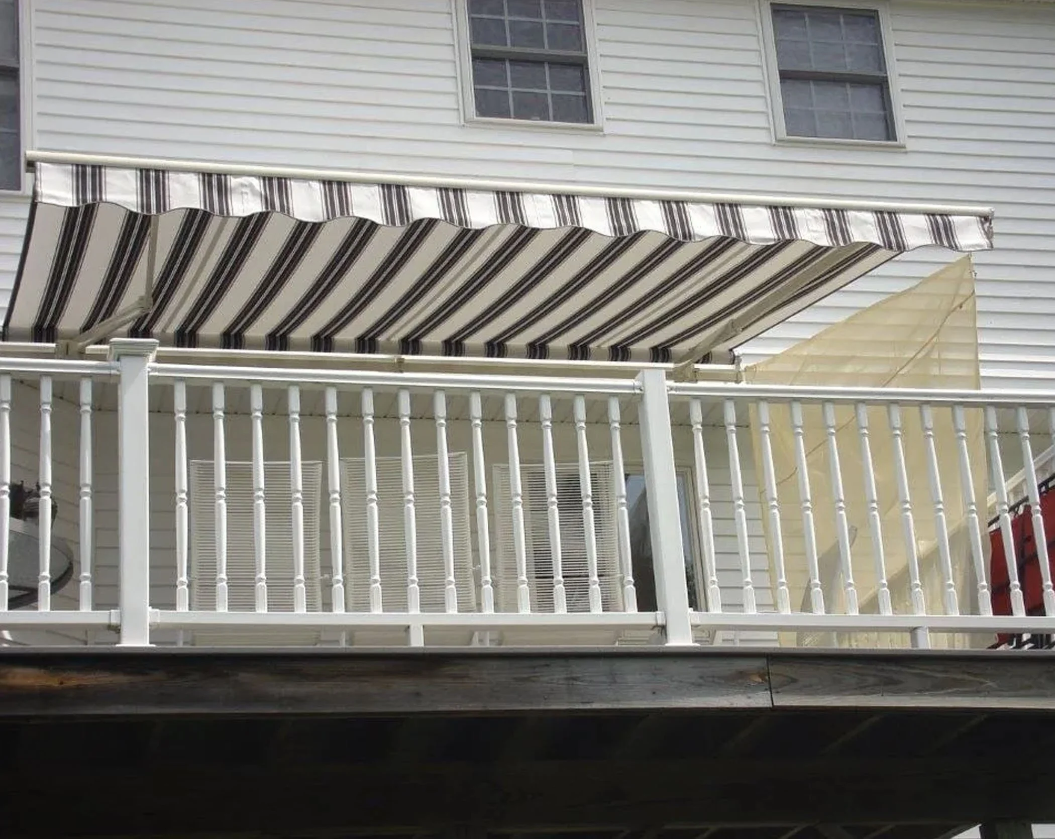 Second-story porch with white railing, striped retractable awning, and beige privacy screen, attached to a white house with double-hung windows.