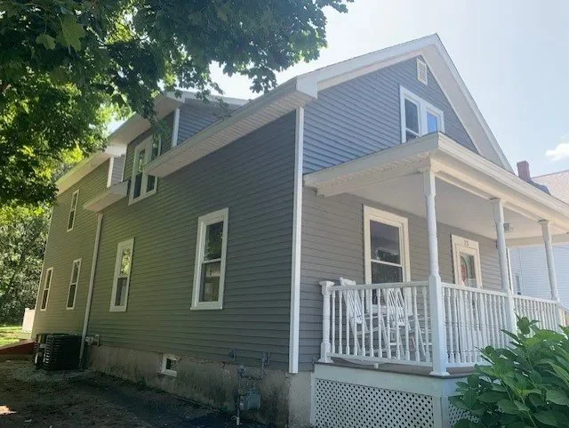 Side view of a gray two-story house with a front porch, white railing, and multiple windows, surrounded by greenery.