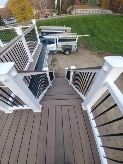 View looking down a set of wooden stairs with white railings onto a backyard with a truck parked on dirt and a grassy lawn.