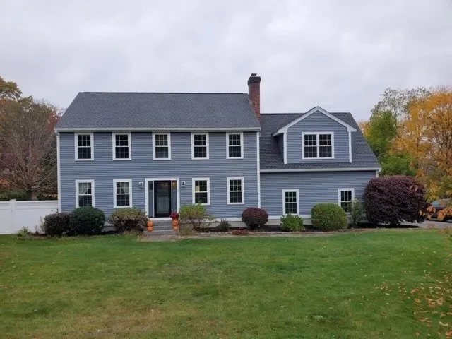 Large blue two-story house with white trim, surrounded by a green lawn and bushes, under an overcast sky.