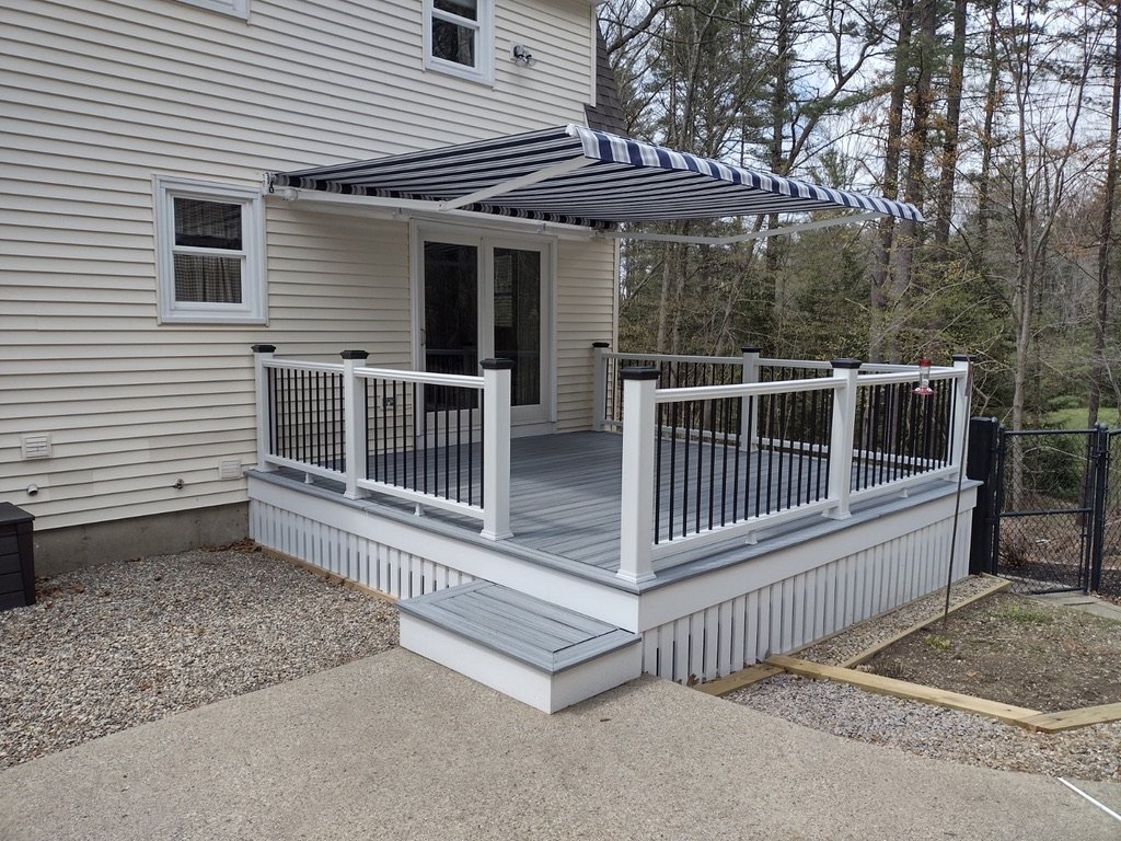 A patio with a raised deck attached to a house, featuring a striped awning and outdoor railing, surrounded by trees and fencing.