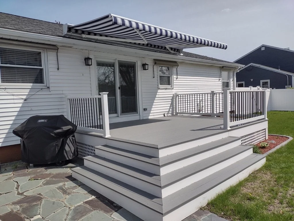 Backyard patio with stairs, railing, barbecue grill, sliding glass door, window, and retractable striped awning on a white house.
