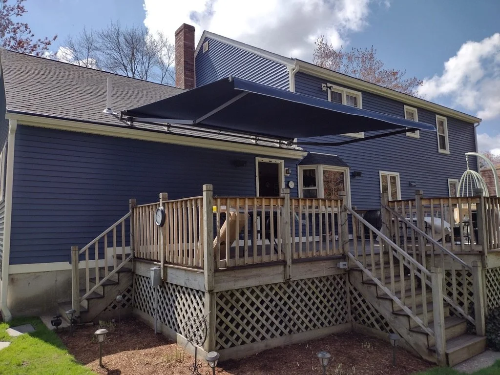 Backyard view of a blue two-story house with a wooden deck, steps, and a black retractable awning, under a partly cloudy sky.