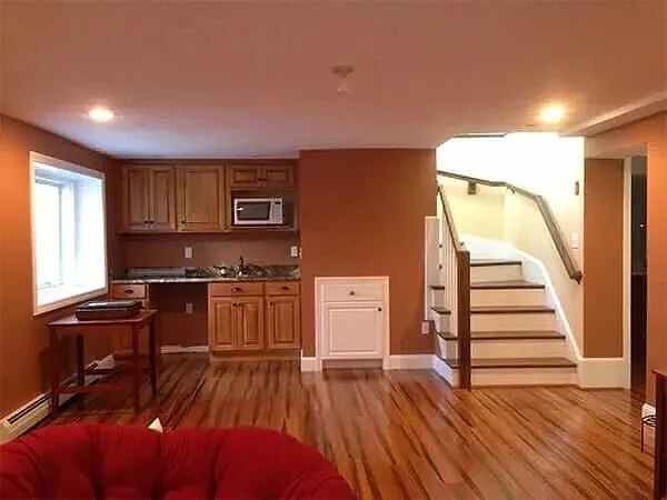 Living room with wooden flooring, a red couch, kitchen area with wooden cabinets, microwave, and small table, and a staircase leading upstairs.