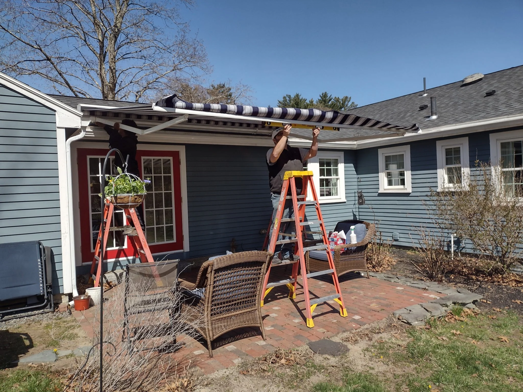 Two people working on installing or repairing a striped awning outside a blue house with a red door and several windows, using yellow ladders under a clear blue sky.