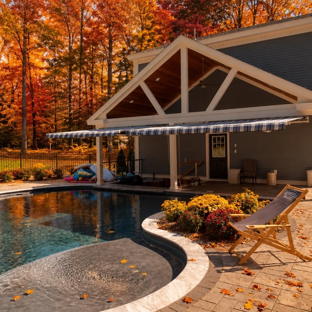 A backyard with a pool and a house with a covered patio. There are autumn leaves on the ground and in the pool, and fall-colored trees in the background.