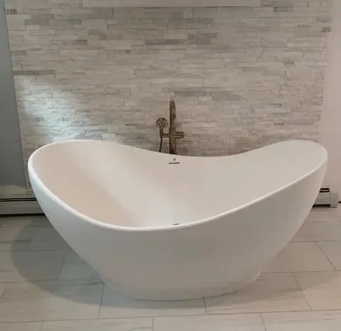 Modern white freestanding bathtub in a bathroom with light gray stone wall and floor tiles.
