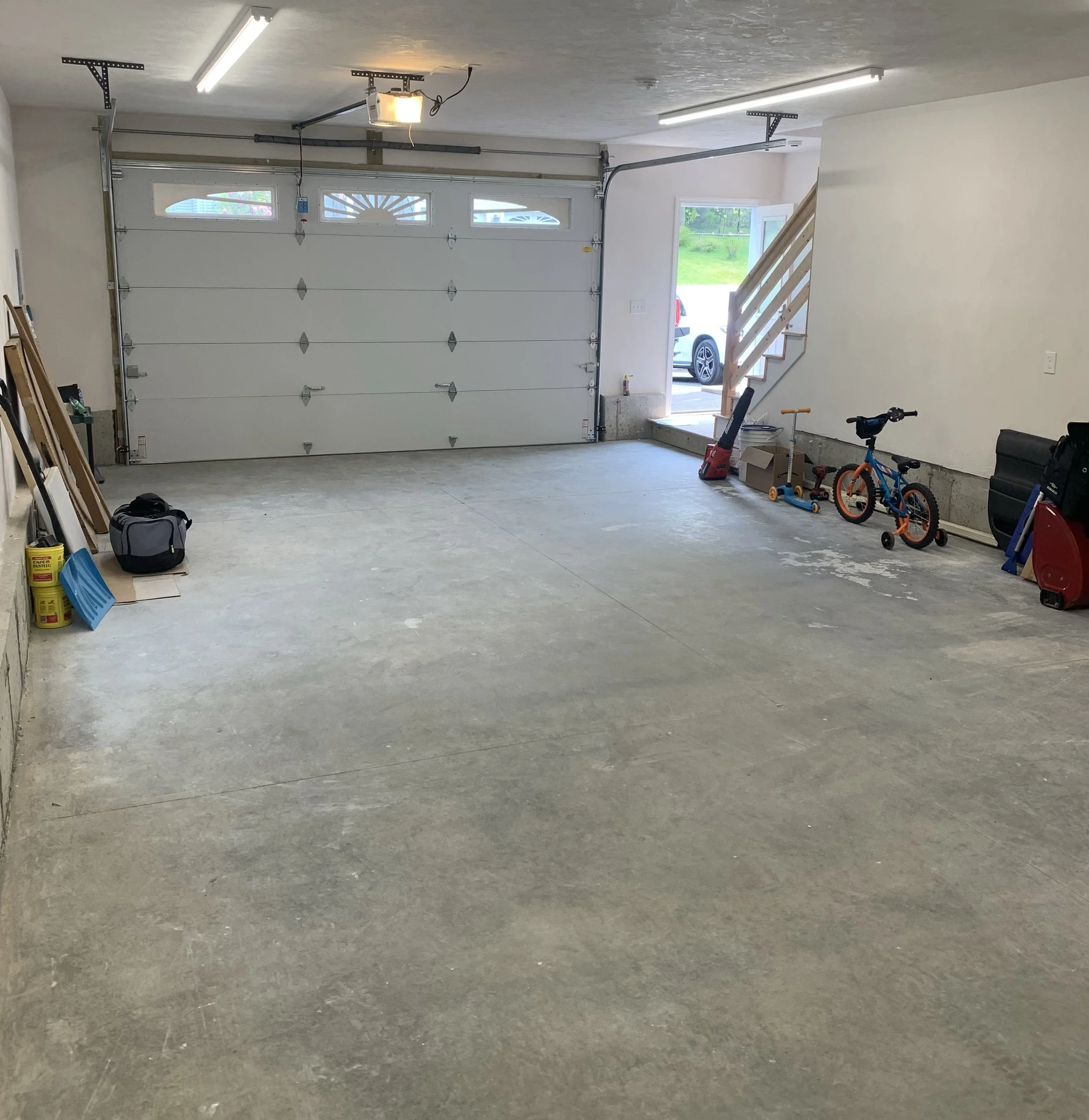 Empty garage with a white sectional garage door, a wooden staircase leading outside, and children's bicycles along the right wall. Various tools and storage items are on the left side.