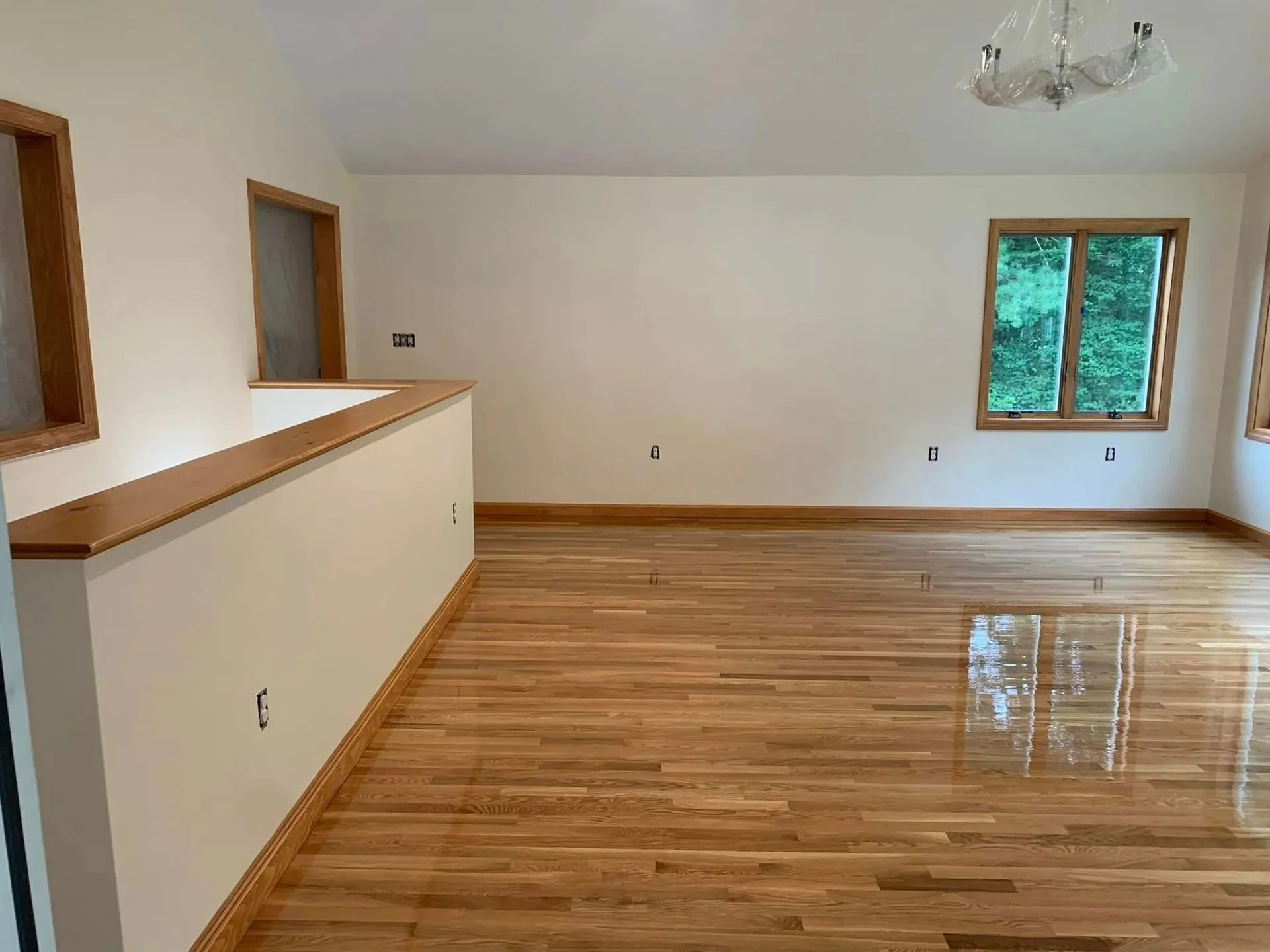Empty living room with polished hardwood floors, white walls, multiple windows, and a modern light fixture on the ceiling.