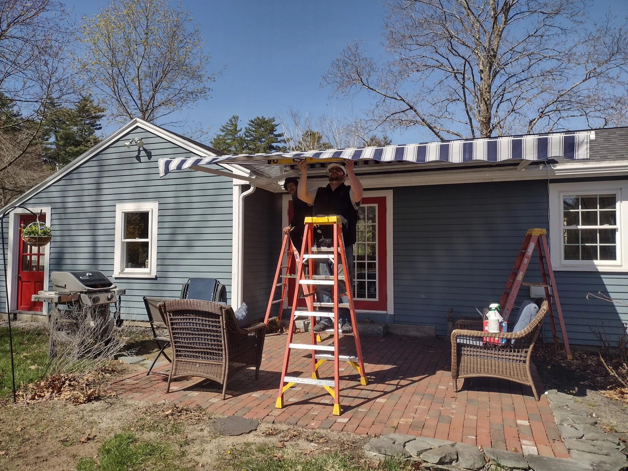 Two men are on a ladder adjusting a striped awning on the back porch of a blue house with red trim. The patio has outdoor furniture and tools, and leafless trees are visible in the background under a clear blue sky.