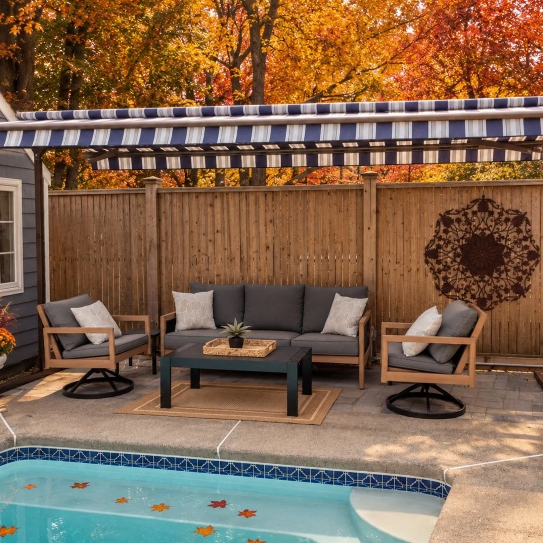 Outdoor patio with a swimming pool, cushioned seating, a coffee table with a plant, fall foliage in the background, and a wooden privacy fence.