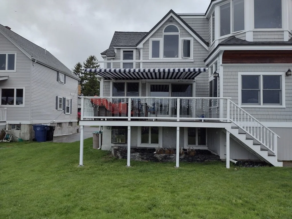 Backyard with a gray house featuring a raised deck with a blue and white striped awning, outdoor furniture, and a set of stairs leading down to a grassy lawn.