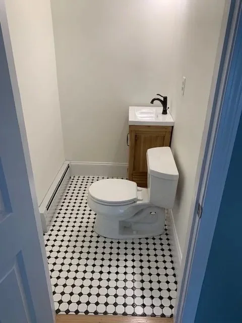 Small bathroom with a white toilet, a small wooden cabinet with a sink and black faucet, and white walls with a black and white tiled floor.
