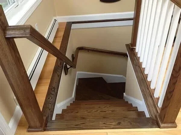 A staircase viewed from the top, showing wooden steps and railings with white spindles, and a view of the lower level with dark hardwood flooring.
