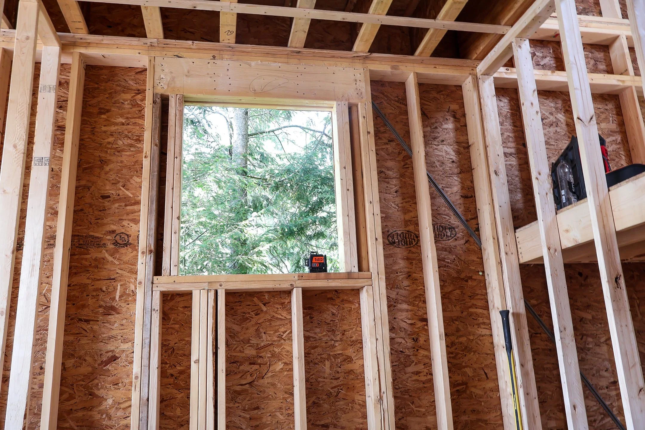 The interior wooden framing of a building under construction with a window opening showing trees outside, construction tools and materials visible.
