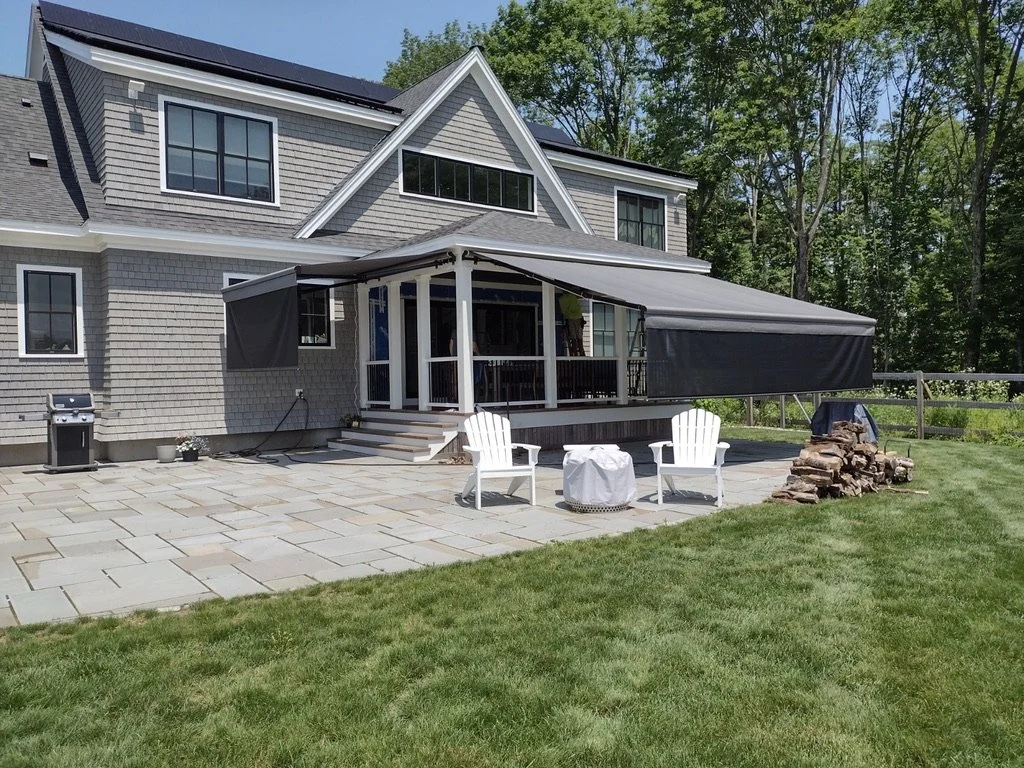 Backyard patio with a house in the background, featuring a screened porch with an extended awning, two white Adirondack chairs, a small table, a woodpile, and a barbecue grill, surrounded by greenery.