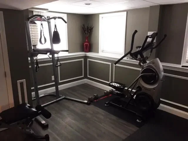 A small home gym with a power rack, boxing bag, rowing machine, and an exercise bench, with a window and decorative vase in the background.