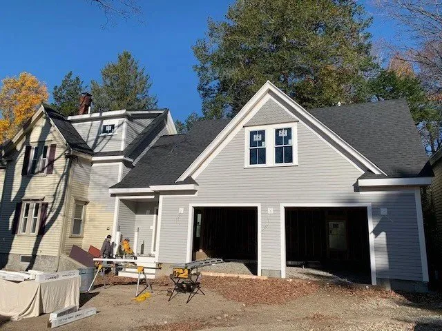 A house under construction with two garage doors, siding, windows, and a pitched roof, set outdoors on a clear day with trees in the background.