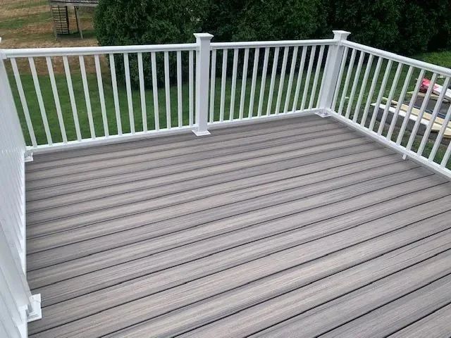 Empty outdoor deck with white railing and gray wooden floorboards, overlooking a grassy backyard with trees.