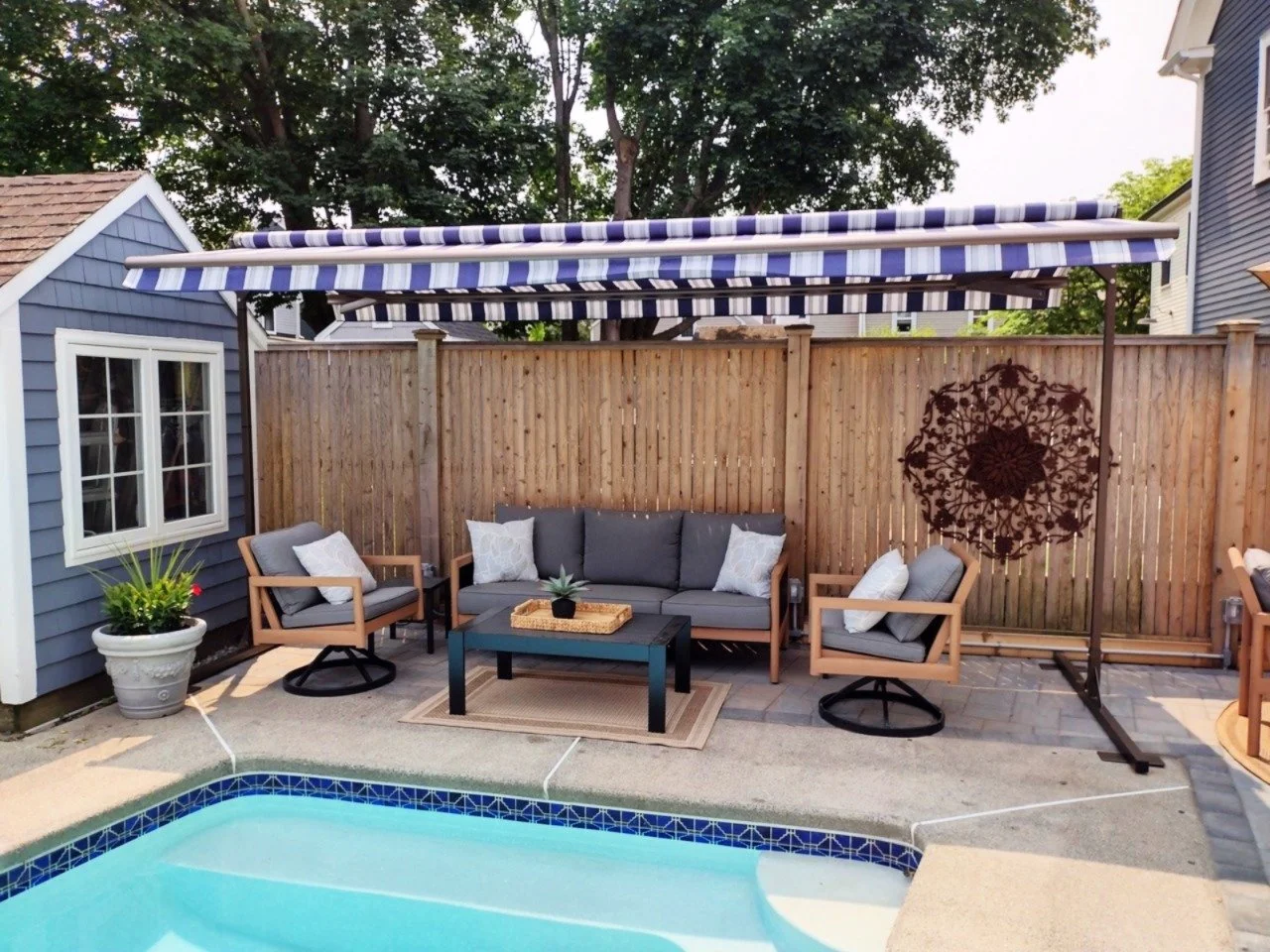 Patio area with a swimming pool, outdoor seating including two chairs and a sofa with white pillows, a small table with a tray and plant, a potted plant, a large striped blue and white awning, a wooden fence, and a decorative sun-shaped metal wall ha