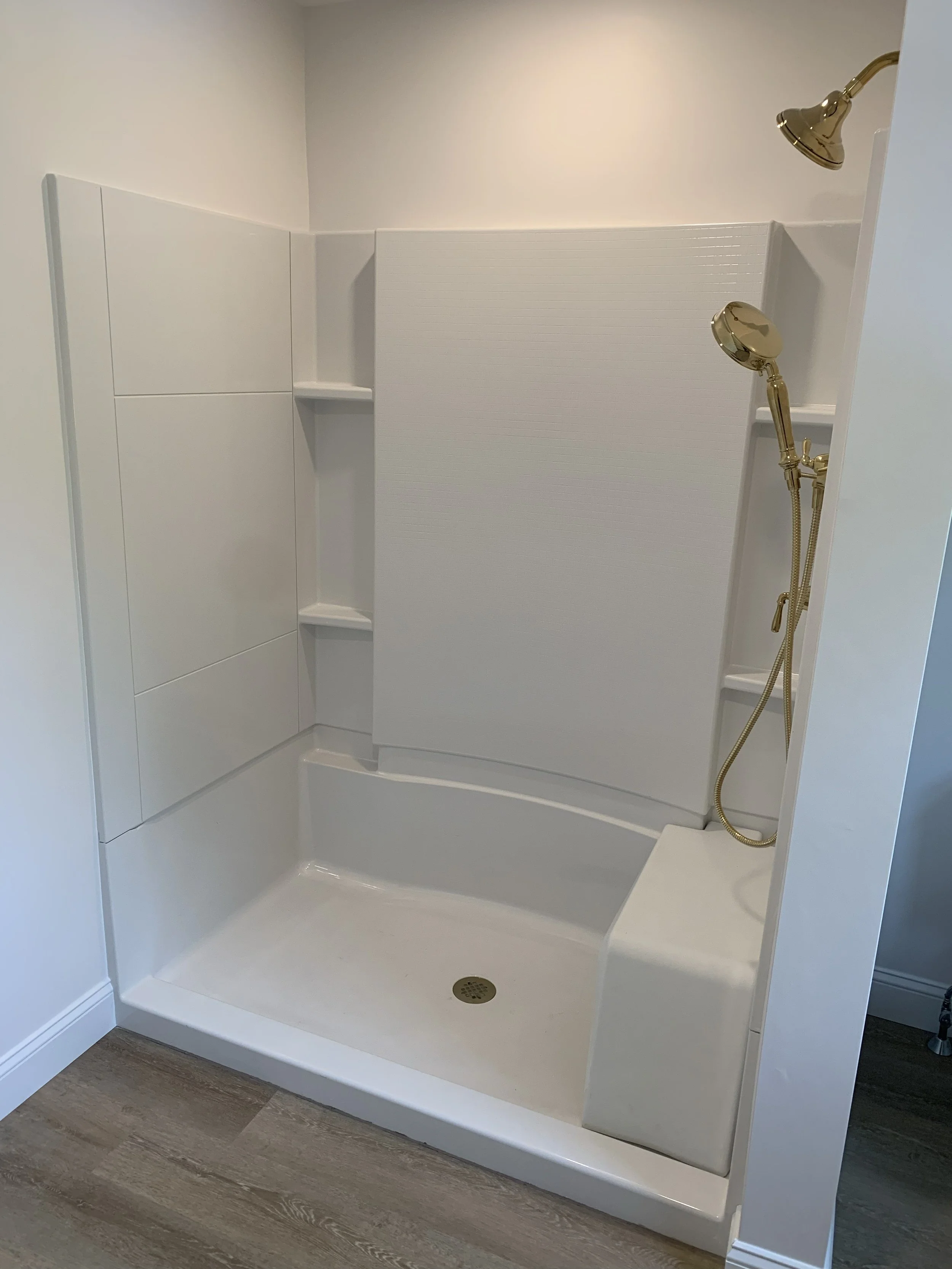Empty white shower stall with built-in shelves, gold shower fixtures, and a textured white divider, on a wood-like floor.