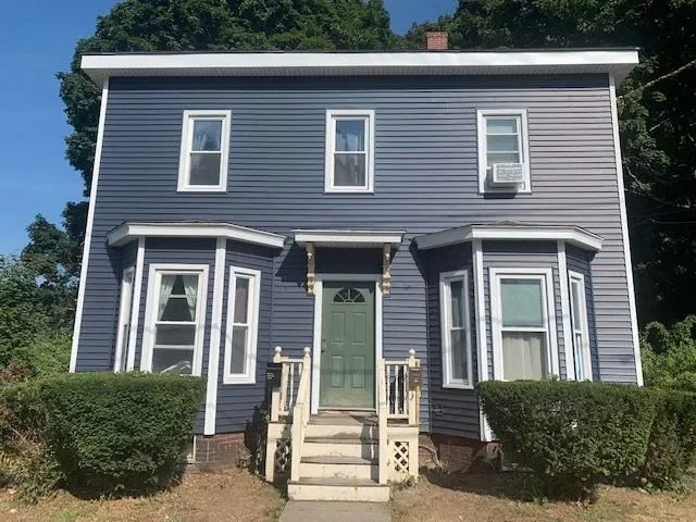 Two-story blue house with four windows on the front, a green front door, and a small front porch with steps and railings. Bushes are on both sides of the stairs, and there are trees in the background.