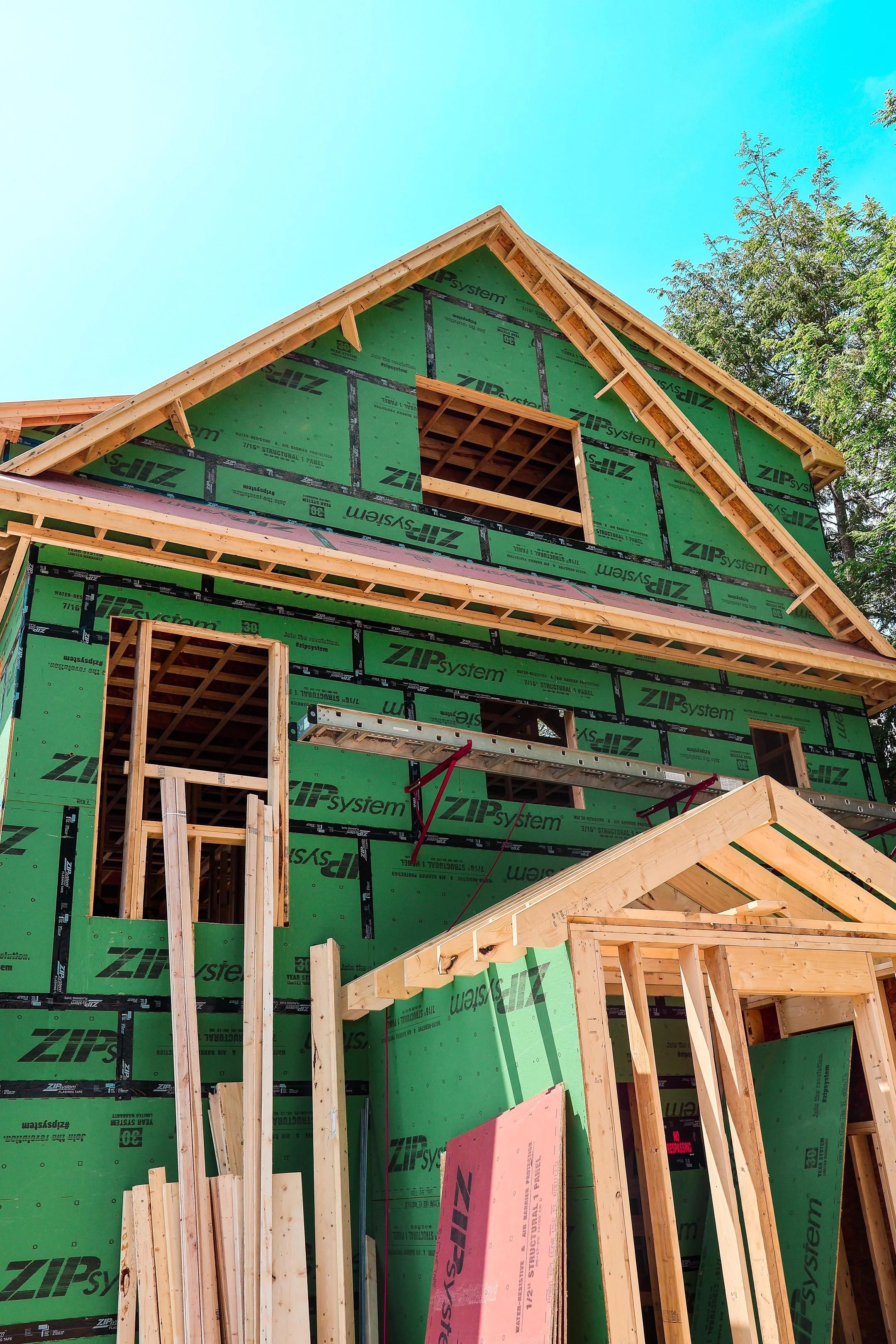 House under construction with green builder's wrap, wooden framing, and a blue sky background.