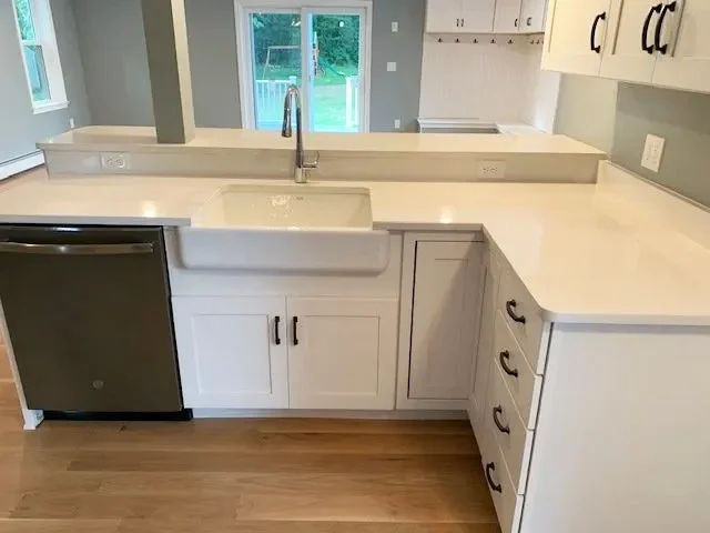 Kitchen with white cabinets, a white farmhouse-style sink, a stainless steel dishwasher, and a long white countertop. There is a window in the background overlooking a deck and green yard.