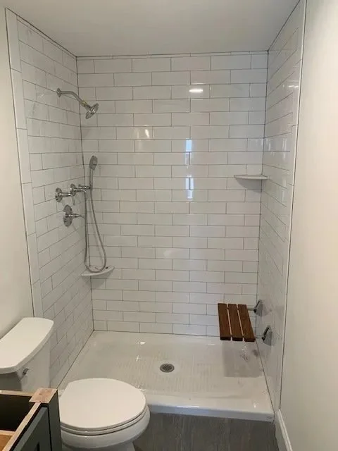 Minimalist bathroom with a walk-in shower featuring white subway tiles, a wall-mounted showerhead, a small wooden bench, and a toilet.