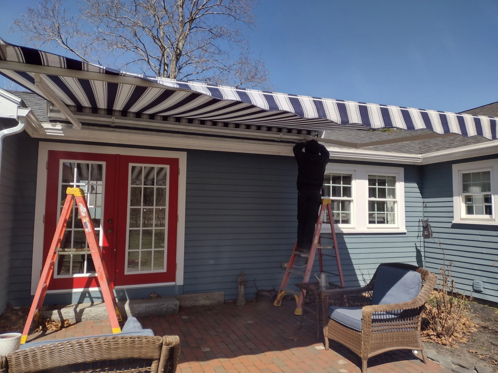 A person installing a striped blue and white retractable awning over a patio area of a house with blue siding and a red door. There are wicker chairs, a small table with a plant, a ladder, and a voice assistant device on the patio. The sky is clear a