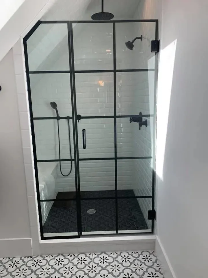 A modern walk-in shower with a black metal frame and clear glass panels, white subway tile walls, a black overhead rain shower, a black handheld showerhead, and a black fixture on the wall, with patterned tile flooring outside the shower.