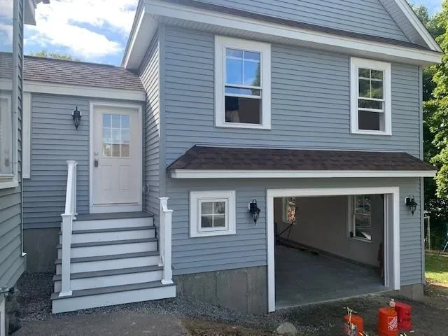 Front of a two-story house with blue siding, white trim, and a garage with an open door. There are stairs leading up to a side door with a small porch area, and several windows on both stories. Outdoor lighting fixtures are visible near the entrance
