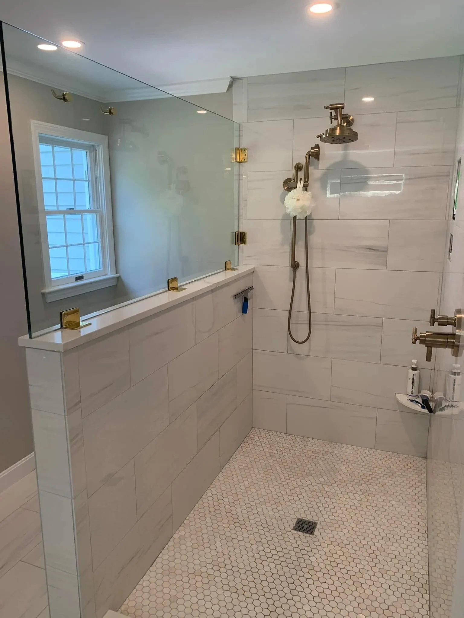 Modern walk-in shower with glass partition, white marble tiles, brass fixtures, showerhead, and a window in a bathroom.